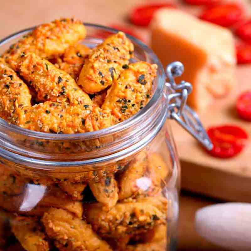 Feinkostgebäck in Glasbehälter, Käse und Tomaten im Hintergrund.