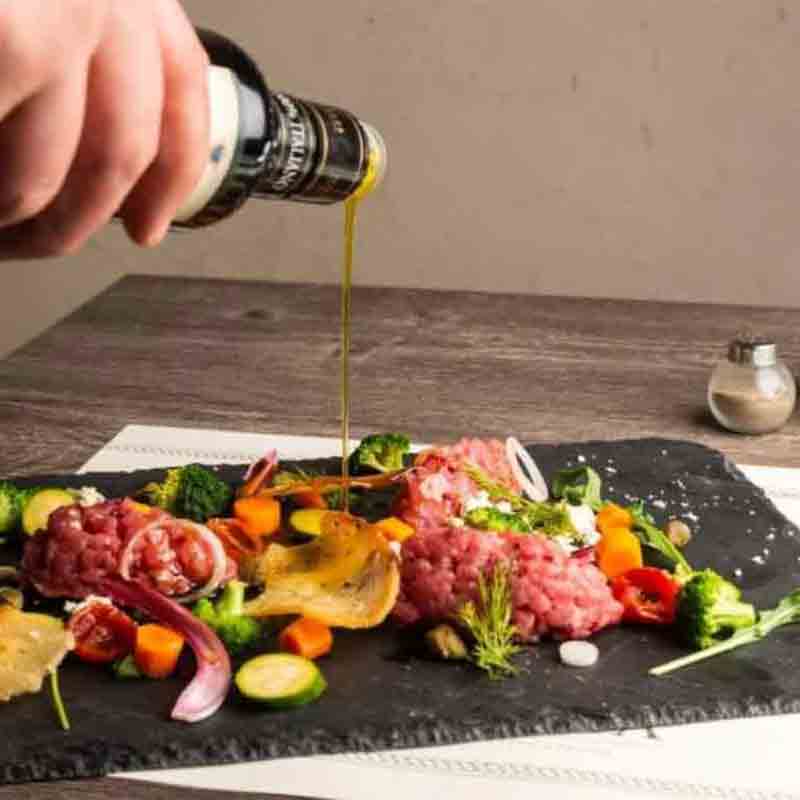 Hand gießt Olivenöl über Teller mit Feinkost und Gemüse.