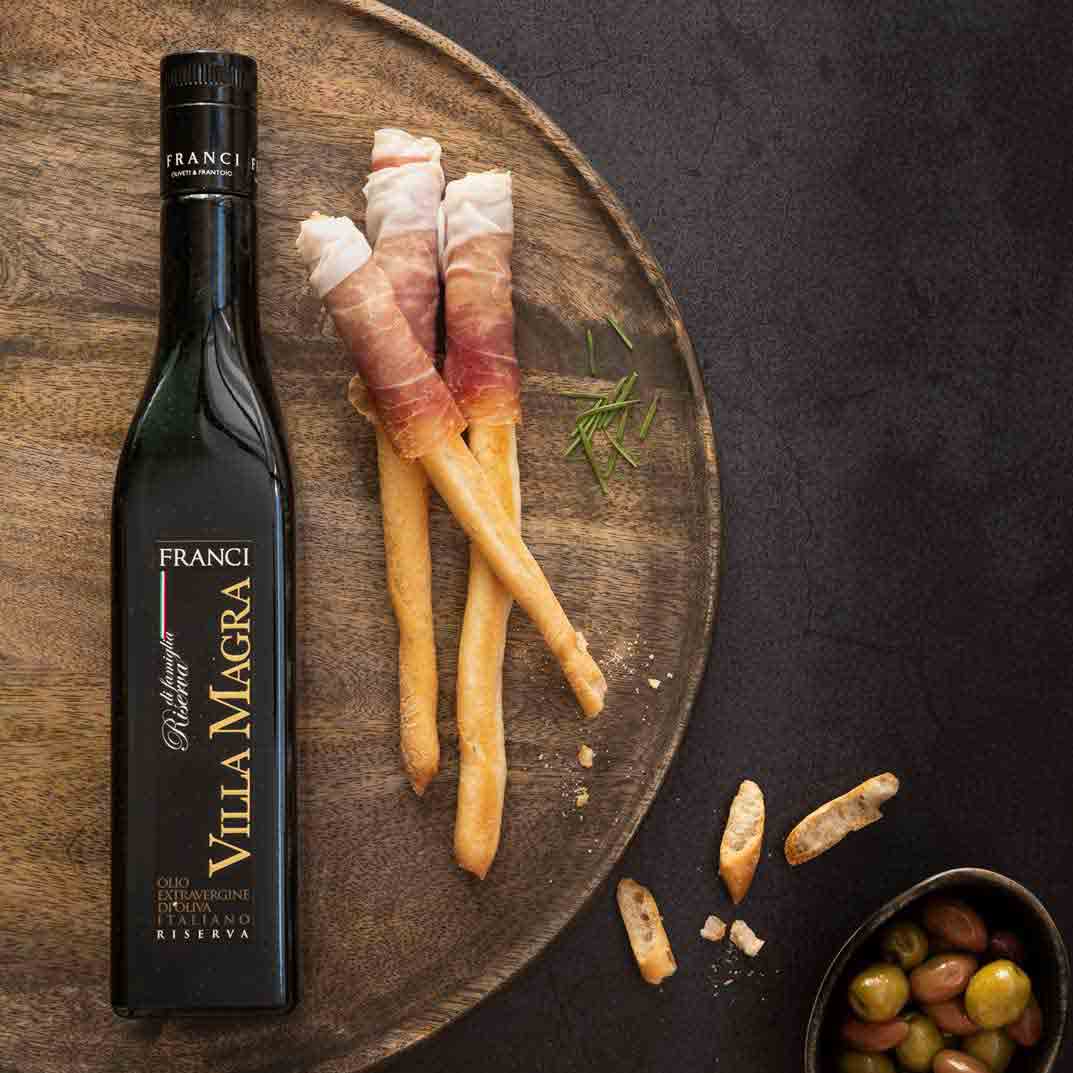 Franci Olivenöl, Grissini, Schinken, Oliven auf Holzplatte.