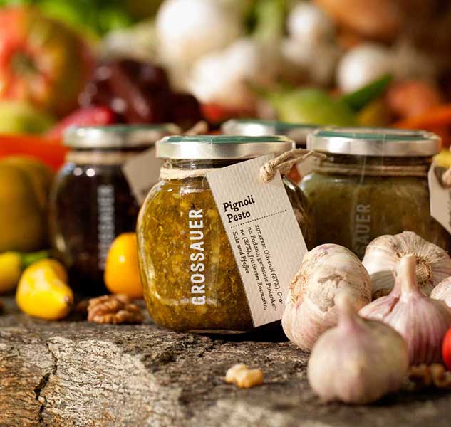 Gläser mit Pesto, Knoblauch, Nüssen und Gemüse im Hintergrund.