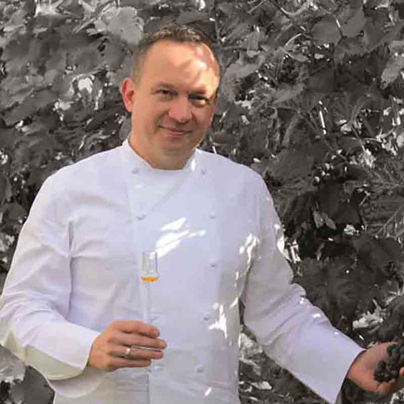 Jens Lang in einer weißen Kochjacke hält ein Glas Aperitifessig und Trauben vor einem monochromen Blattwerk.