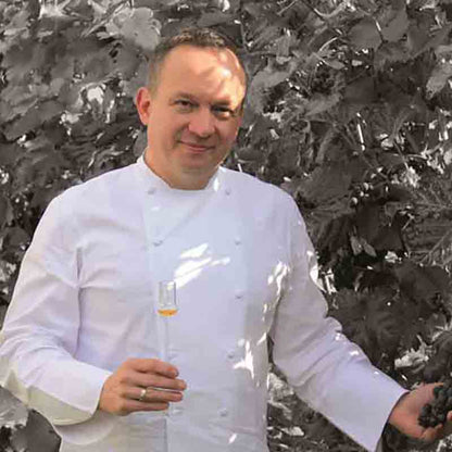 Jens Lang in einer weißen Kochjacke hält ein Glas Aperitifessig und Trauben vor einem monochromen Blattwerk.