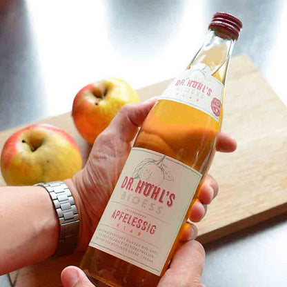 Person hält Dr. Höhl's BioEss Apfelessig vor Äpfeln.