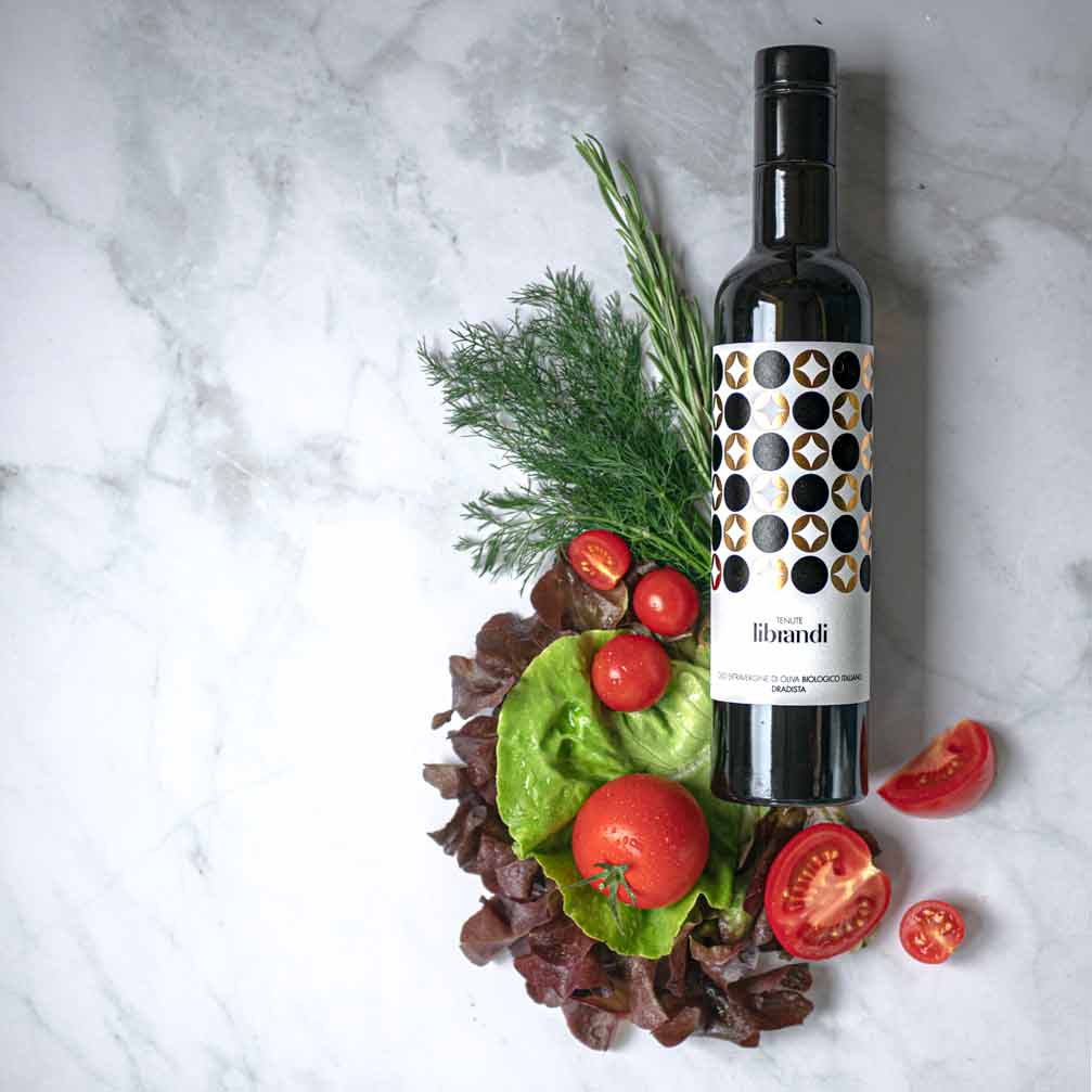 Librandi Olivenölflasche mit frischem Gemüse auf Marmortisch.