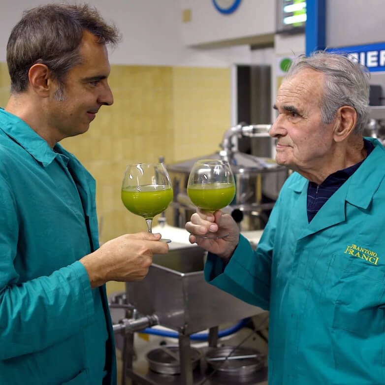 Zwei Männer stoßen mit Gläsern grünem Olivenöl an.
