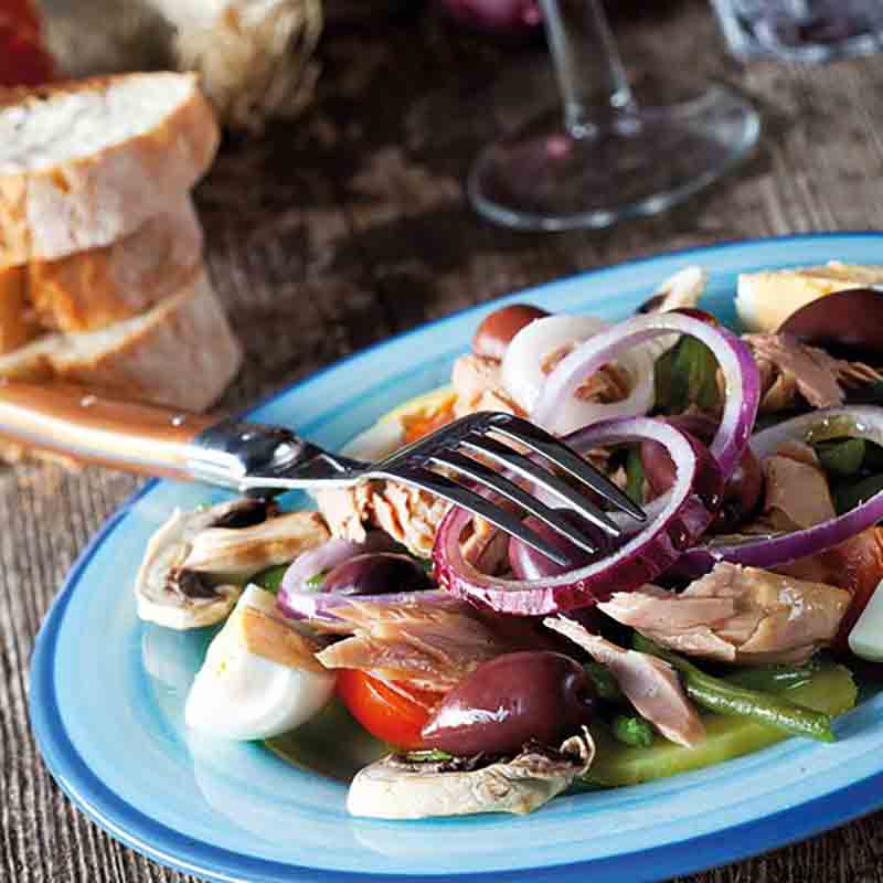 Thunfischsalat mit Oliven, Zwiebeln, Tomaten und Brot im Hintergrund.
