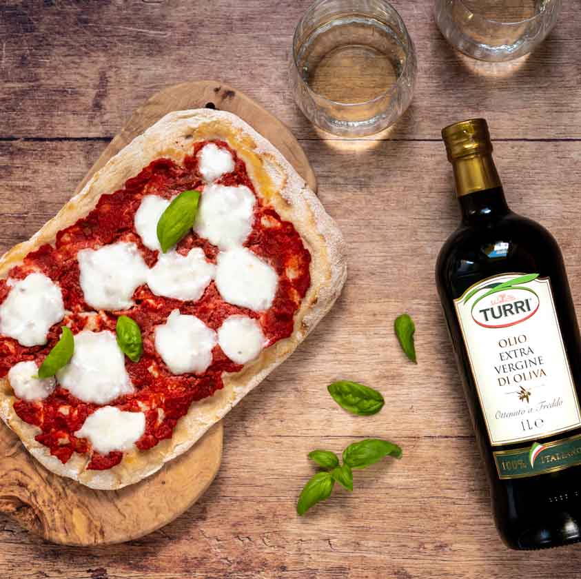 Pizza mit Mozzarella, Basilikum und Turri Olivenölflasche auf Holztisch.