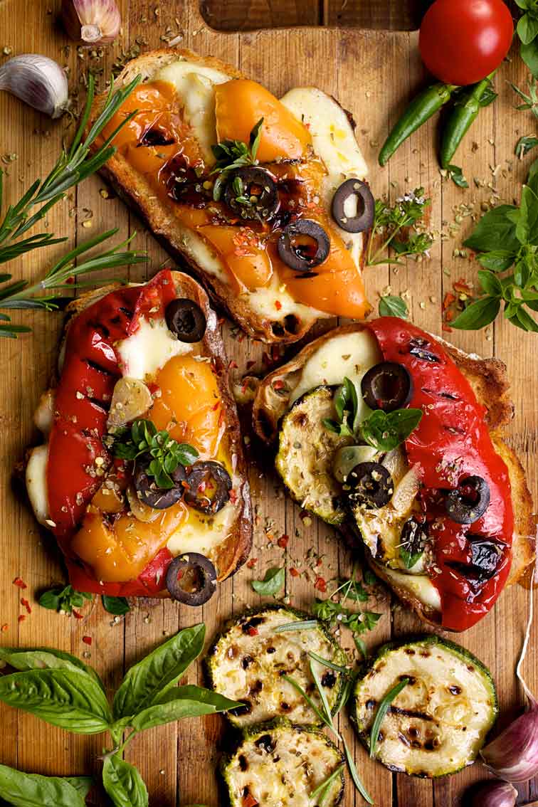 Bruschetta mit Käse, Paprika, Oliven und Kräutern auf Holzbrett.