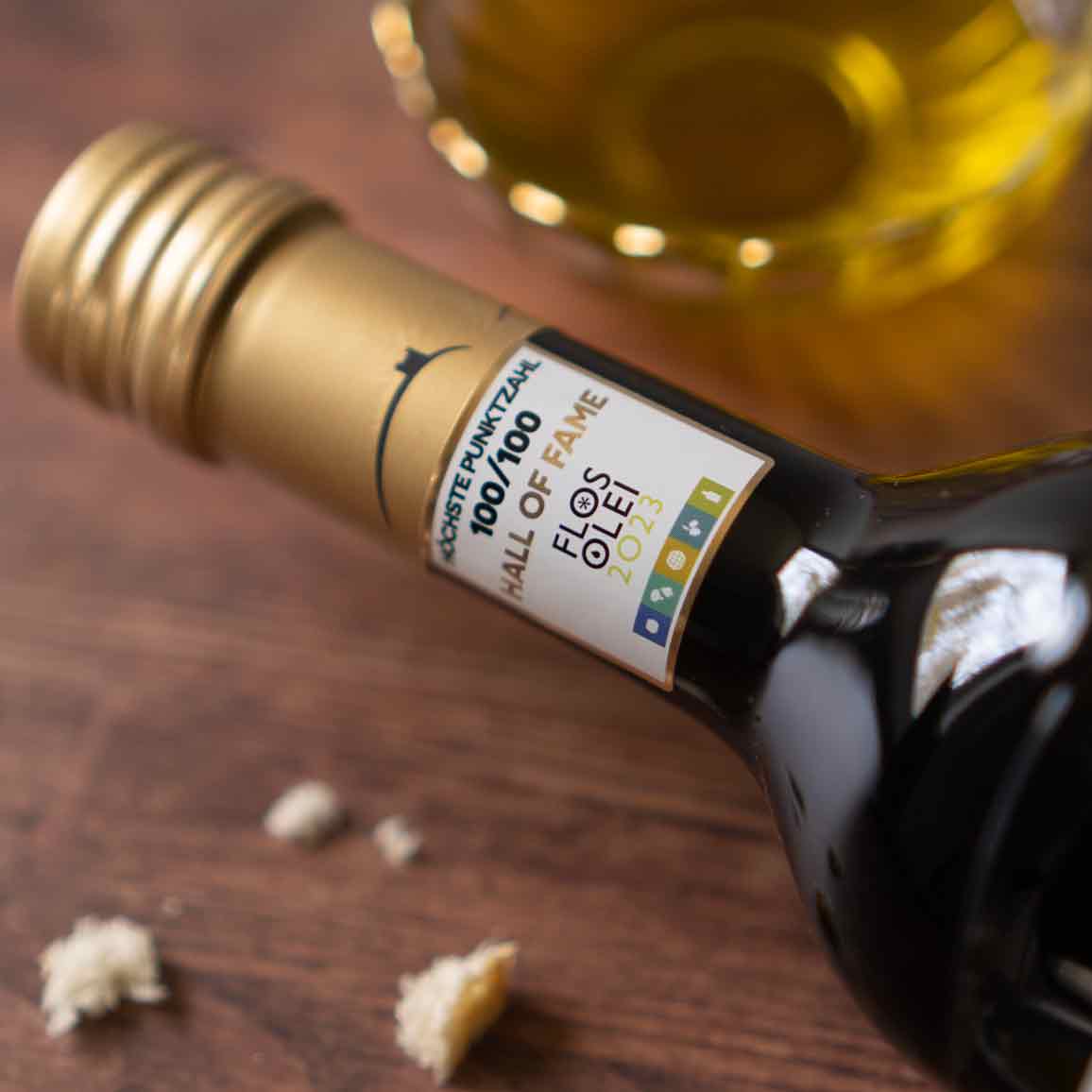 Flos Olei Olivenöl-Flasche auf Holztisch mit Brotkrümeln.