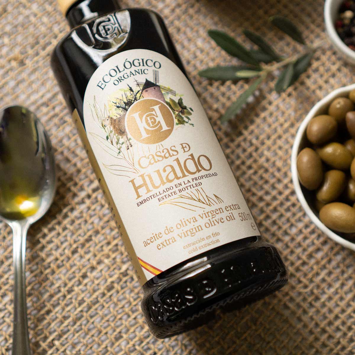 Casas de Hualdo Olivenölflasche neben Oliven und Löffel.