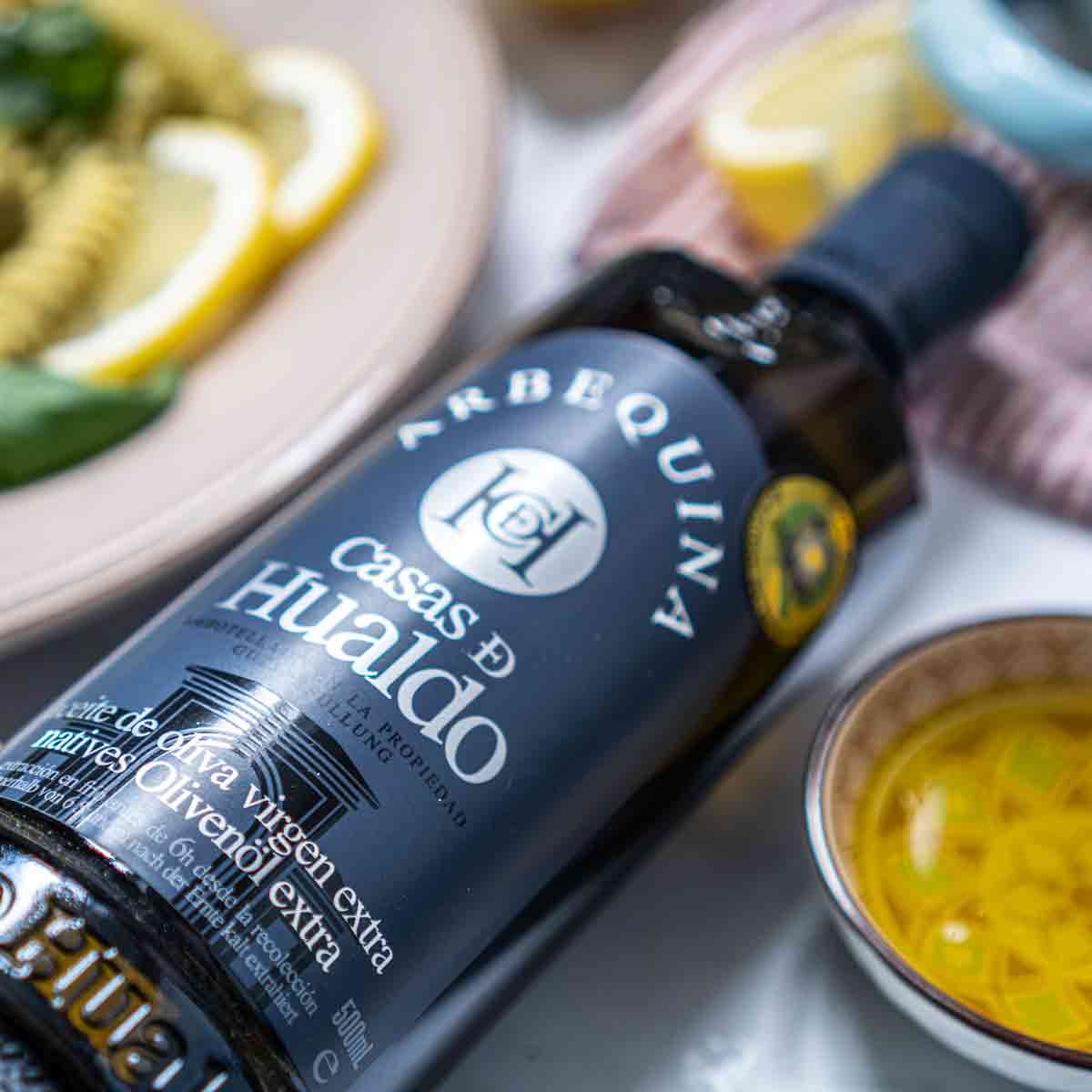 Olivenölflasche Casas de Hualdo neben Speisen und Schalen.