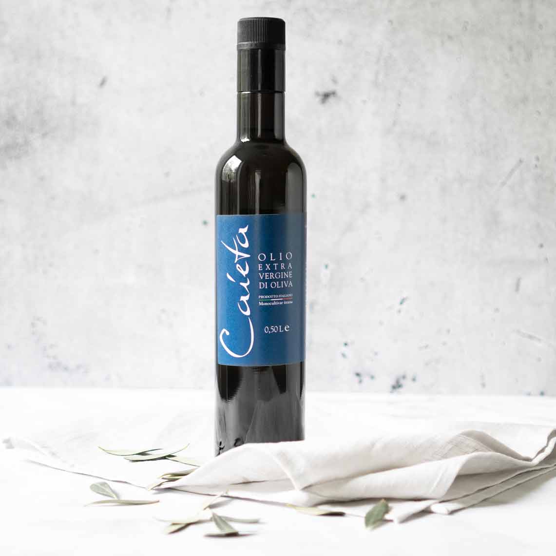 Olivenölflasche Caieta auf weißem Tuch mit Olivenblättern.