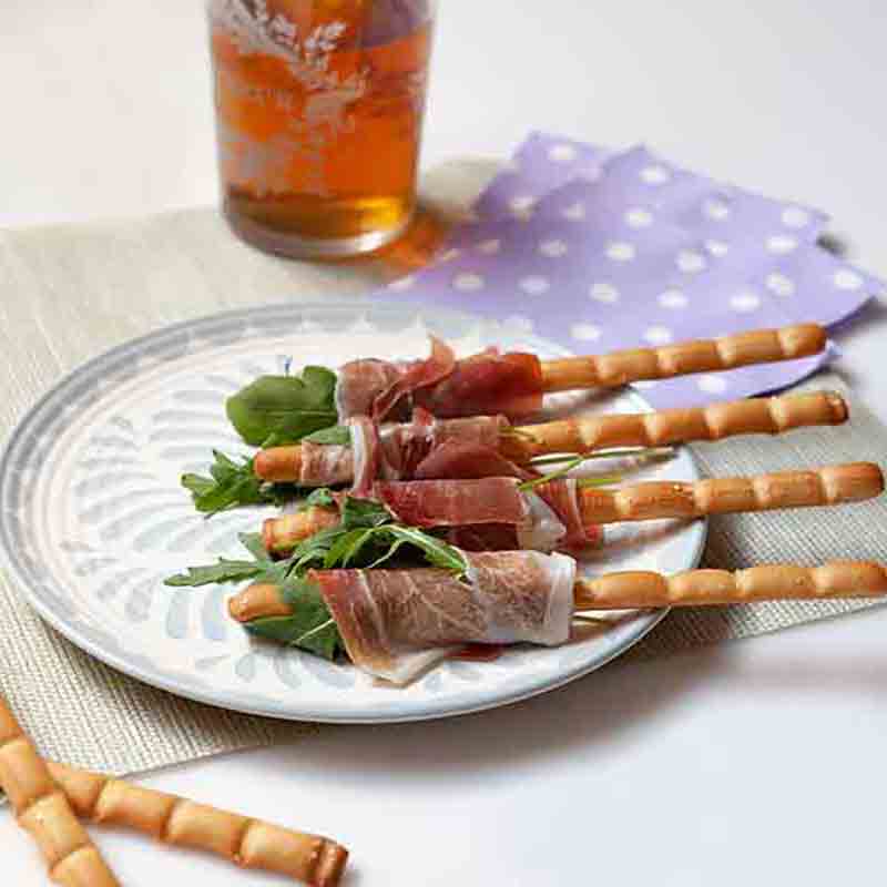 Grissini mit Schinken und Rucola auf Teller, Getränk daneben.