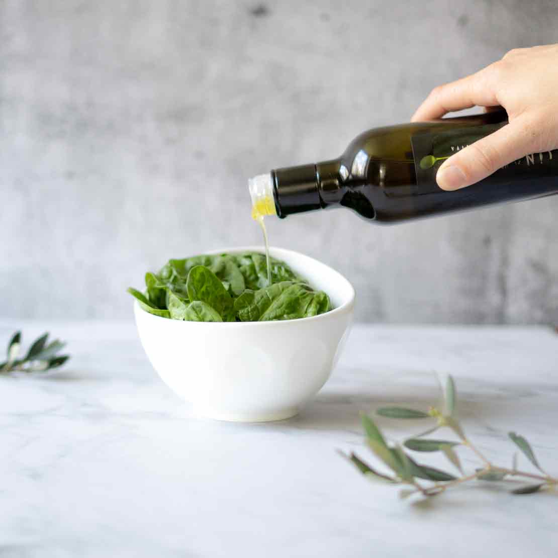 Hand gießt Olivenöl über Salat in weißer Schüssel.