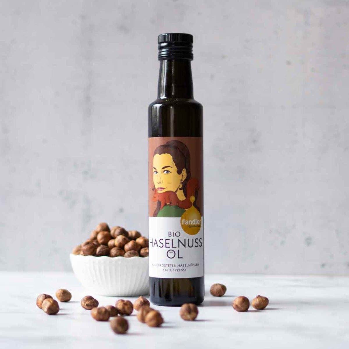 Fandler Bio Haselnussöl Flasche mit Haselnüssen im Hintergrund.