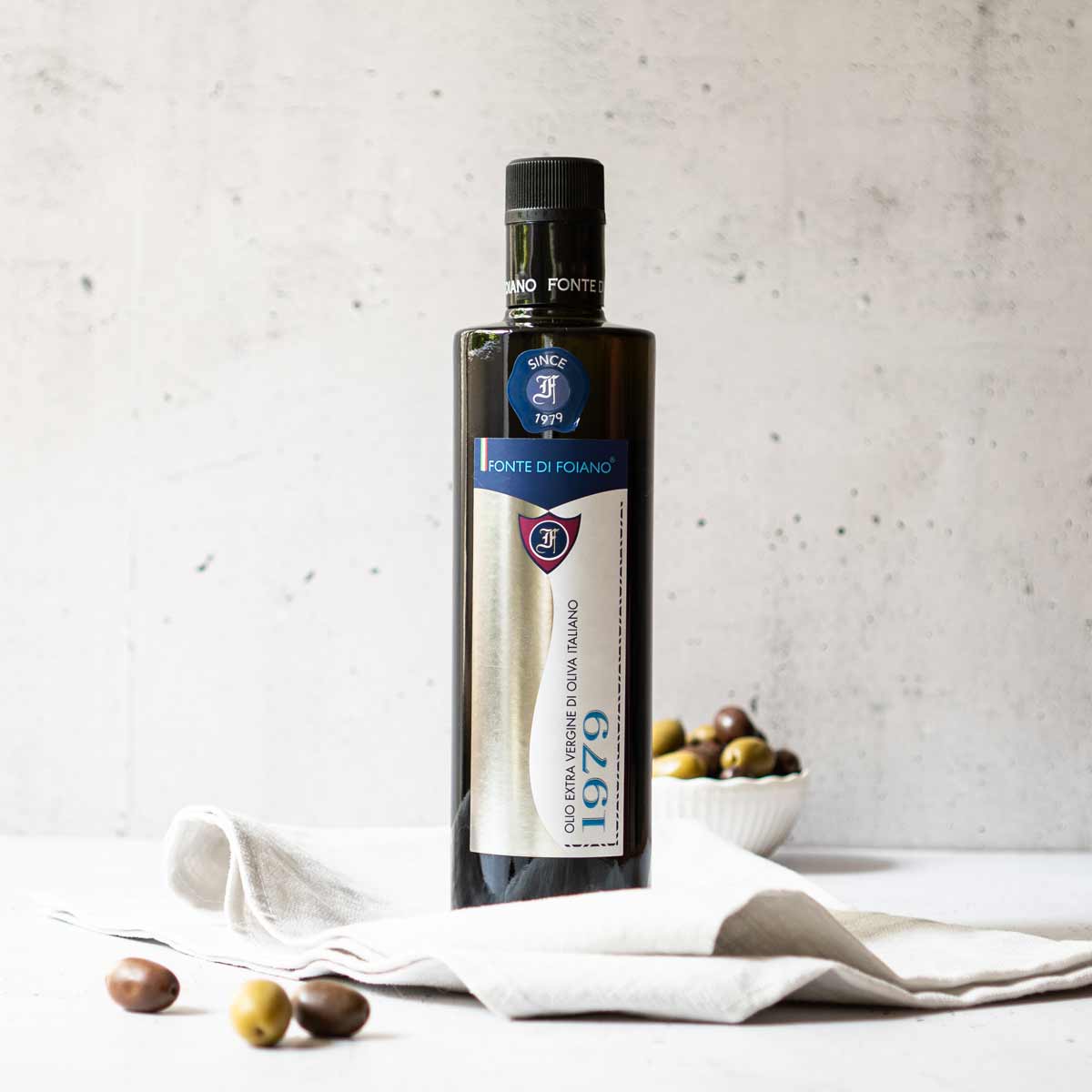 Olivenölflasche Fonte di Foiano mit Oliven auf Tuch.