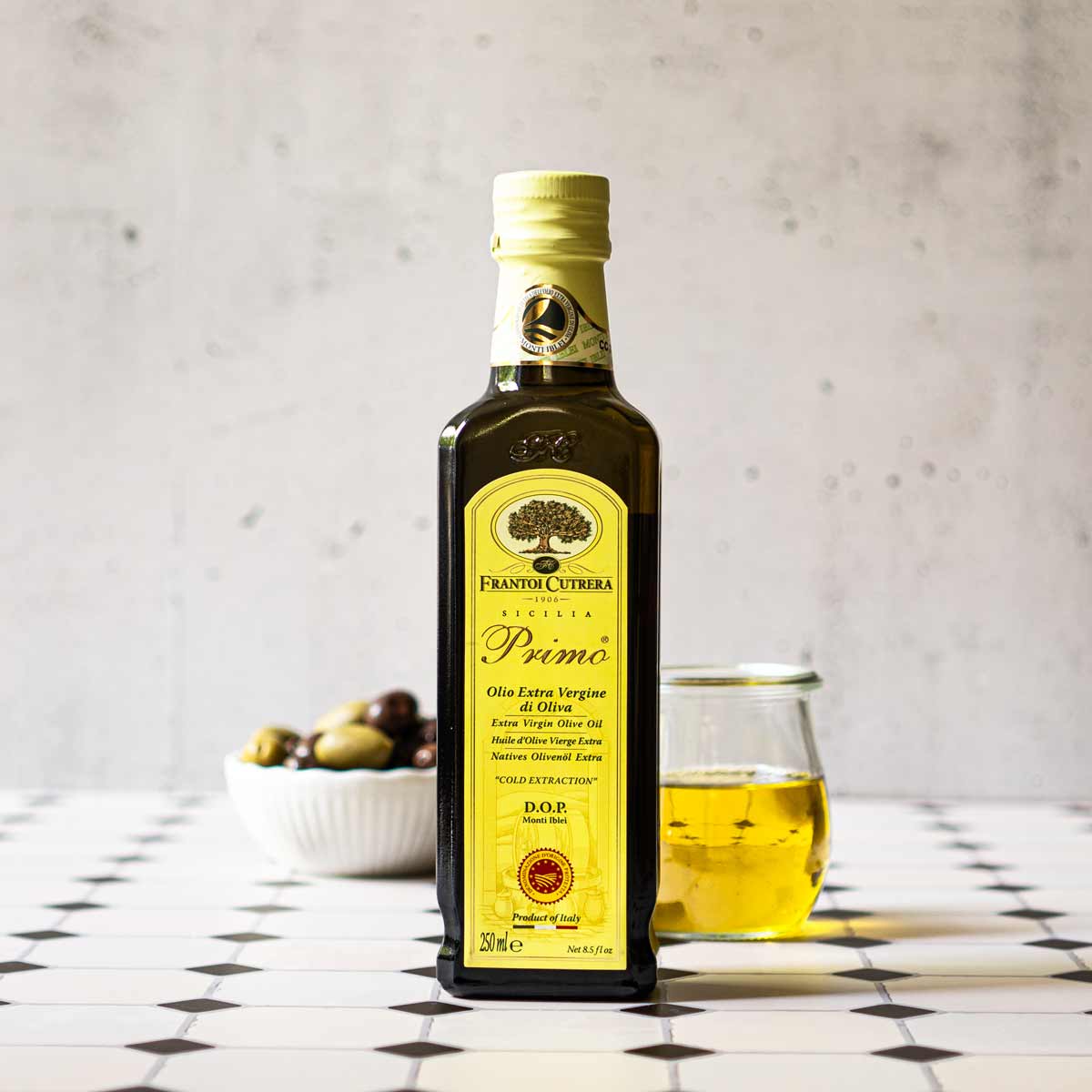 Frantoi Cutrera Primo Olivenölflasche mit Oliven und Glas.