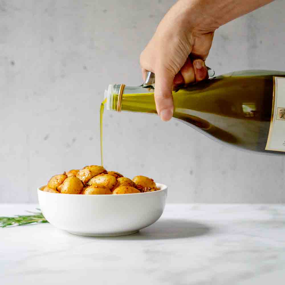 Hand gießt Olivenöl über Kartoffeln in weißer Schüssel.