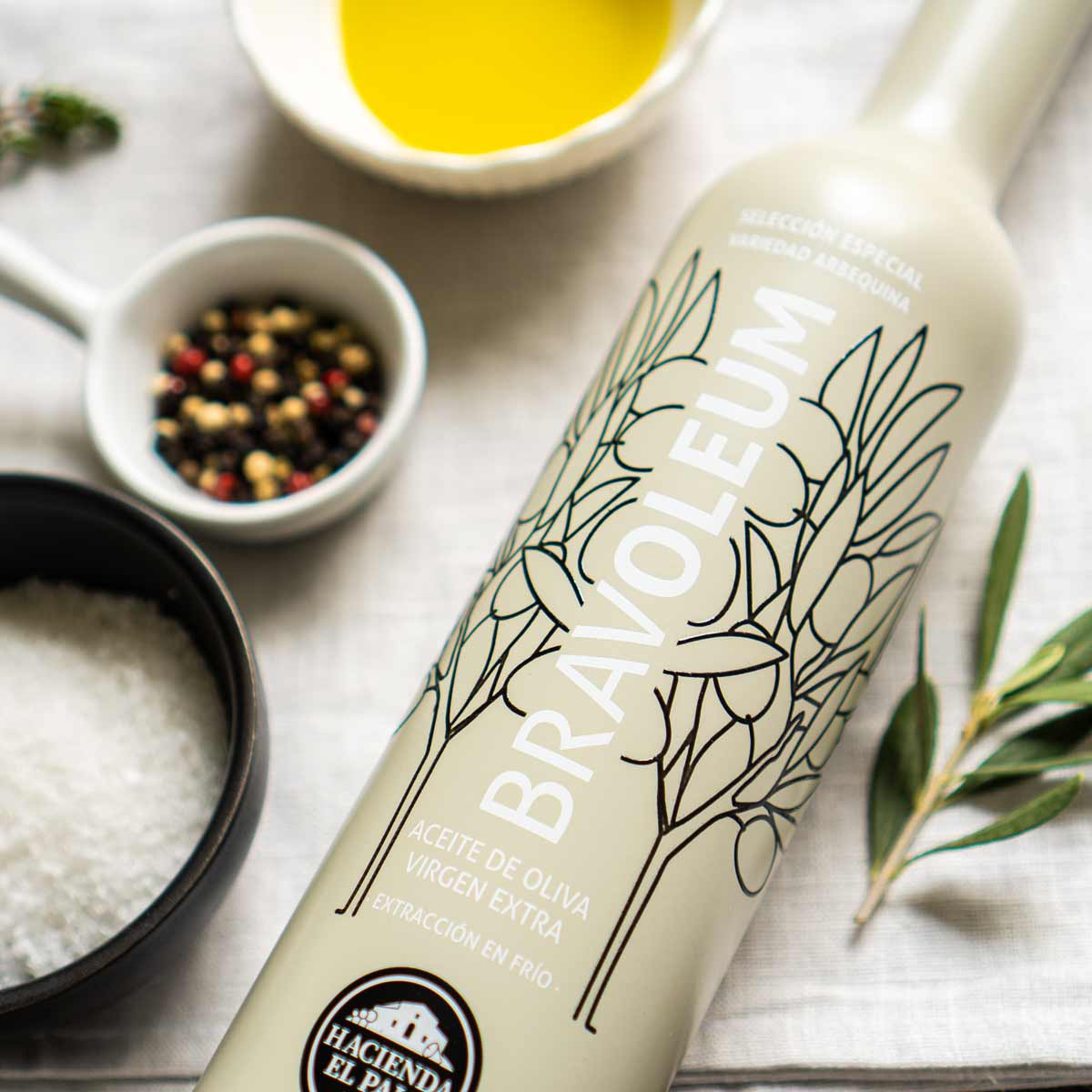 Bravoleum Olivenölflasche mit Gewürzen und Olivenöl im Hintergrund.