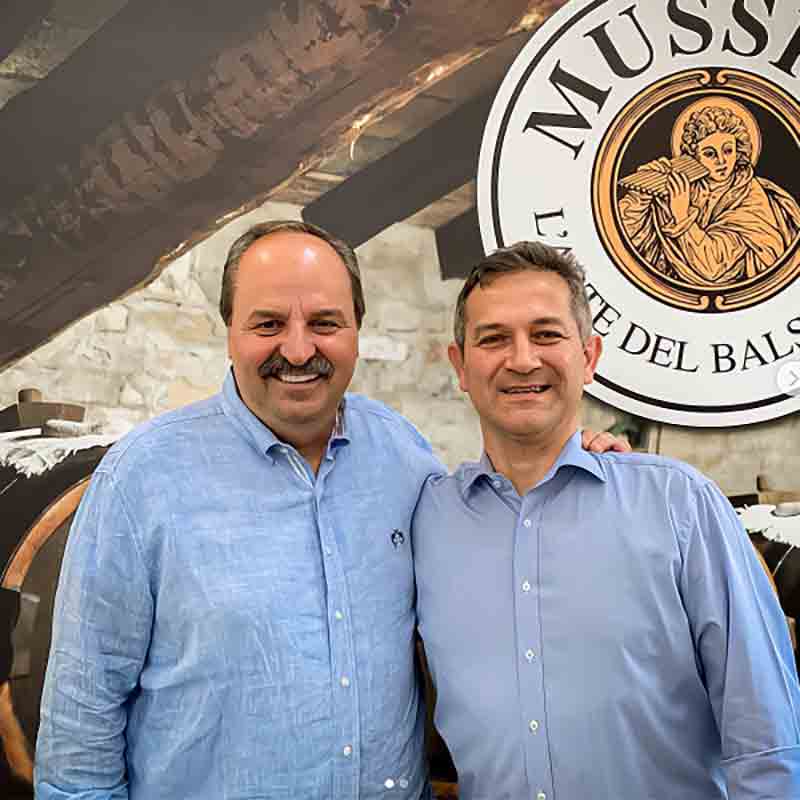 Zwei Männer, beide in hellblauen Hemden, lächeln herzlich vor einem „MUSS“-Logohintergrund mit einer Figur, die einen Stab hält. Ihre Kameradschaft ist so erfrischend wie das Teilen eines Balsam Limoncino – Essigzubereitung für Cocktails von Mussini an einem sonnigen Tag.
