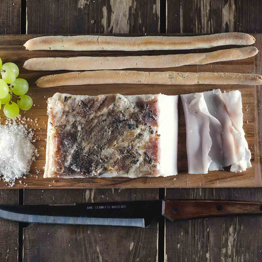 Lardo, Brotstangen, Trauben, Salz und Messer auf Holzbrett.