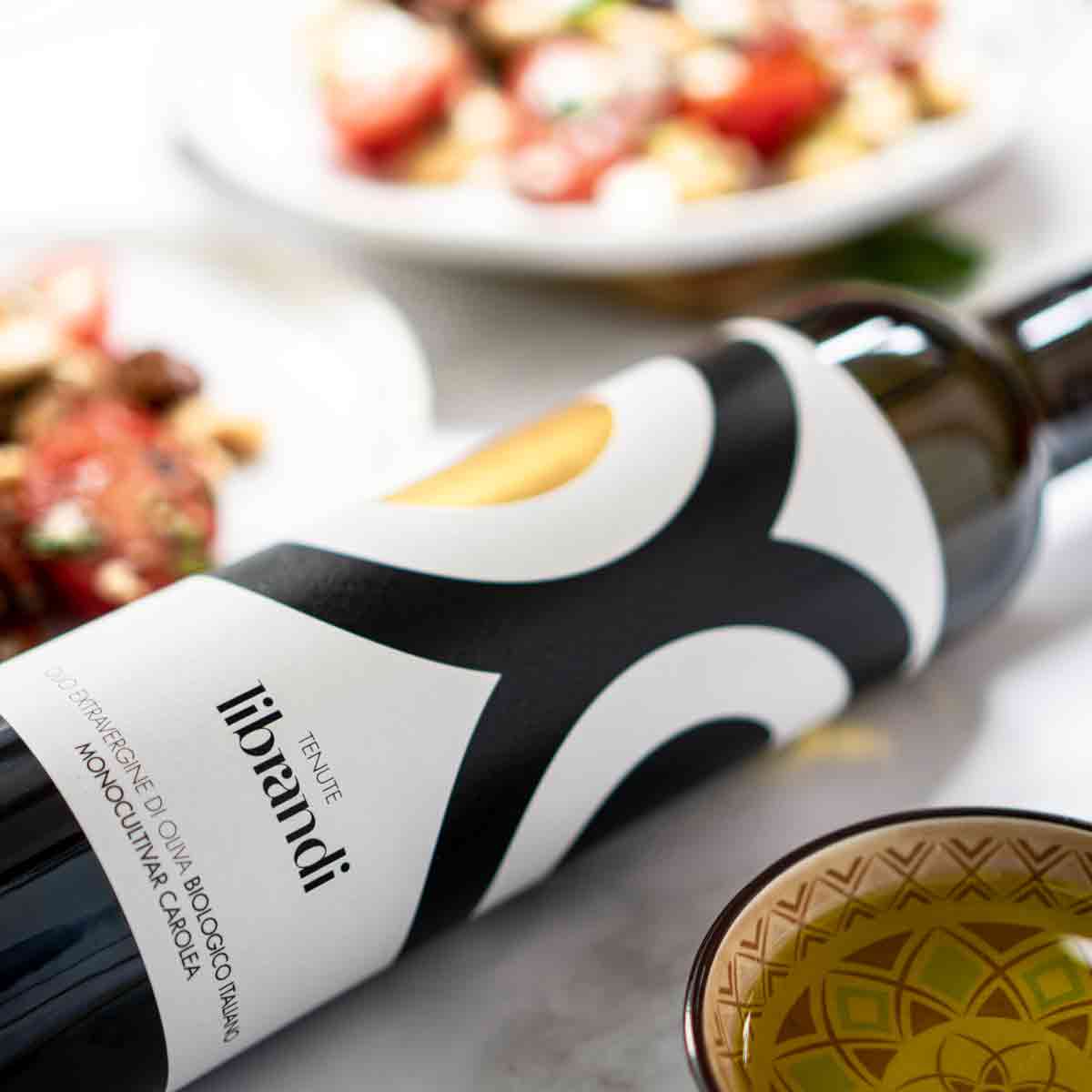 Librandi Olivenölflasche mit Speisen im Hintergrund.