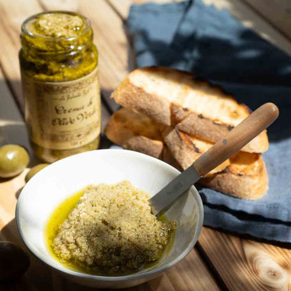 Olivencreme im Glas, Schale, Messer, Brot auf Holztisch.
