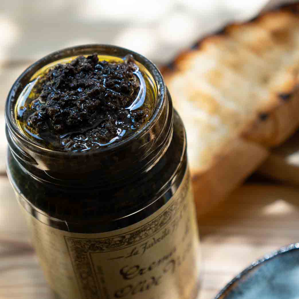 Schwarze Olivencreme im Glas neben geröstetem Brot.