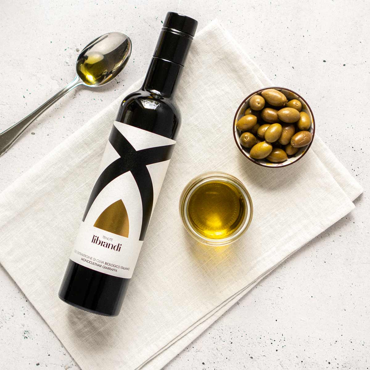 Olivenölflasche Tenute Librandi, Oliven, Löffel, Glas auf Tuch.