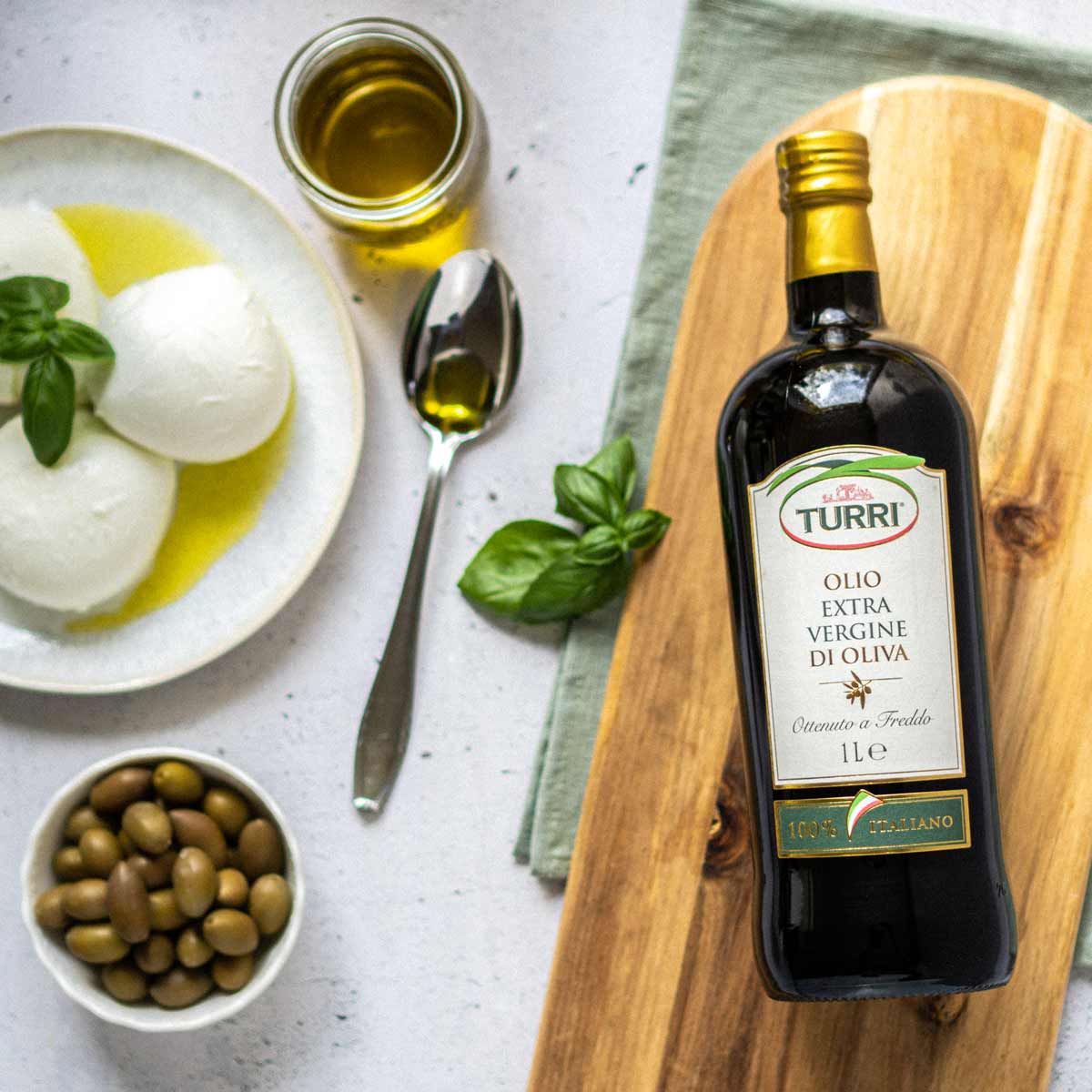 Olivenölflasche Turri, Mozzarella, Oliven, Basilikum, Löffel, Holzbrett.