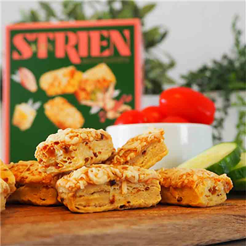 Käsegebäck vor Verpackung mit "Strien" Aufschrift, Gemüse im Hintergrund.